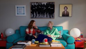 Two women sit on a bright blue couch in a sunny living room.